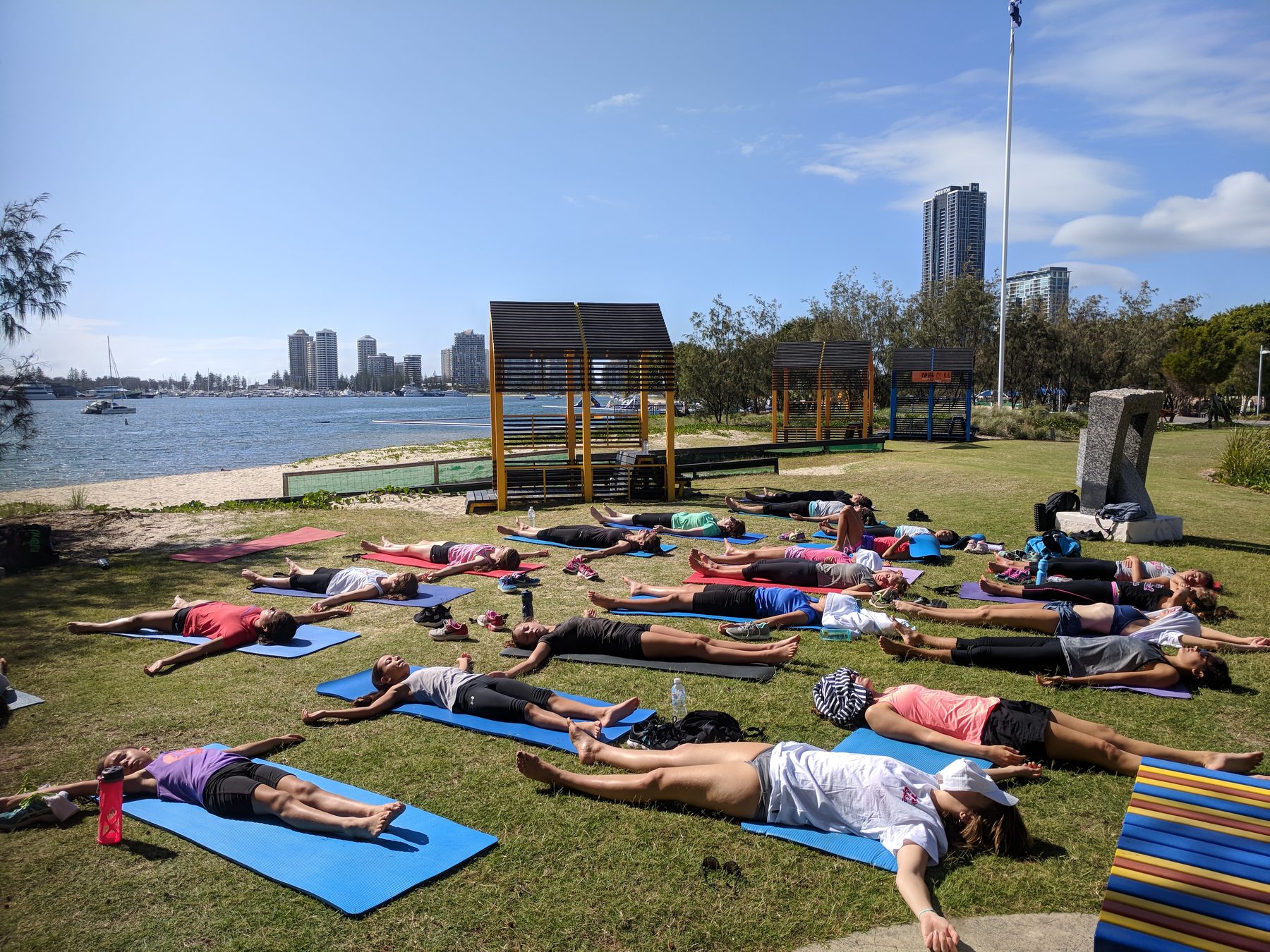 A large group relaxing in savasana on mats at an outdoor waterfront park